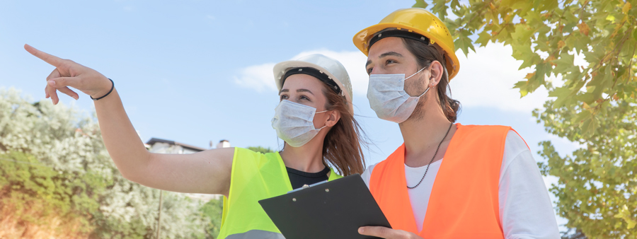 male and female wearing safety vests, safety helmets, and masks looking at something off in the distance. Male holding a clipboard writing information down while female points at something in the distnace.